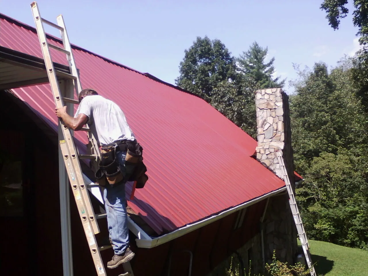 Roofer on a ladder beside a red metal roof and stone chimney — Soffit & Fascia Repair by Roofing Brothers in Pacific